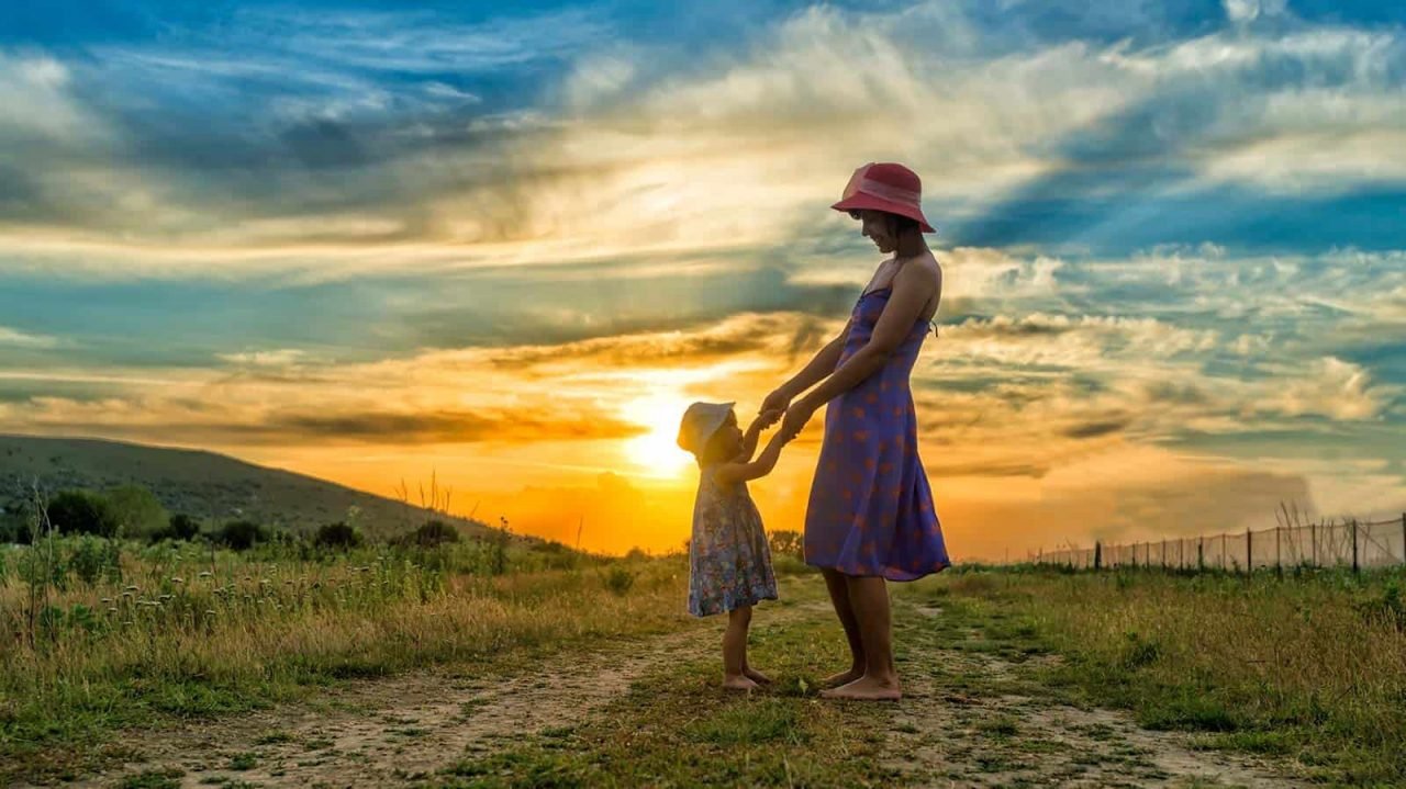 Mãe de chapéu de mãos dadas com a pequena filha num caminho de terra na natureza com por do sol ao fundo
